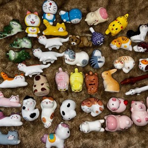 May include: A collection of small ceramic chopstick rests featuring various animal shapes including cats, frogs, rabbits, pigs, and cartoon characters in diverse colours like white, yellow, pink, brown, and blue with painted details.