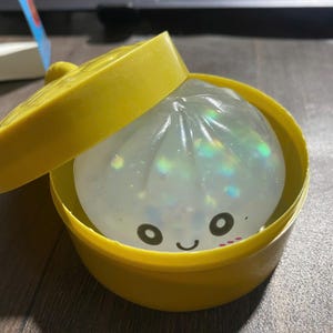 以下が含まれることがあります： 内部に虹色のラメが入った半透明で柔らかい餃子型の玩具。2つの目と笑顔の口が描かれたシンプルな黒い顔がプリントされており、鮮やかな黄色の円形プラスチック容器と蓋の中に収められています。