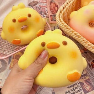 May include: A soft, yellow, chick-shaped squishy toy with a rounded body, small orange beak, brown eyes, and orange feet. The toy has a smooth, matte texture and is being held, showing its pliable nature. Another similar yellow chick toy is visible nearby.