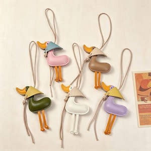 May include: Five duck-shaped leather charms with long thin straps. Each charm features a coloured body, a contrasting yellow or beige beak, and a conical hat. The colours include olive green, pink, brown, white, and lavender, all with orange legs.