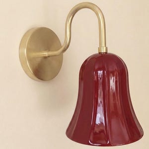May include: A wall-mounted light fixture featuring a curved, brushed brass arm and a circular wall plate. The lamp includes a bell-shaped, deep red glass shade with vertical fluted detailing that extends downward from the brass hardware.