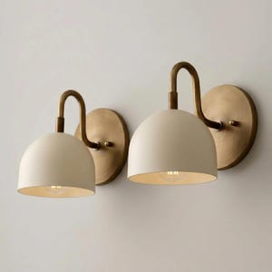 May include: A pair of wall sconces featuring matte cream-coloured dome shades and curved antique brass arms. Each light fixture is mounted on a circular brass backplate, showcasing a clean, functional design with exposed light bulbs inside.