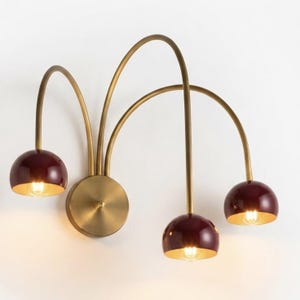 May include: A wall-mounted light fixture featuring a circular brass base with three curved brass arms extending outward. Each arm supports a glossy, deep burgundy dome-shaped shade that houses a single light bulb, emitting a warm glow.