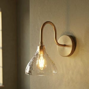 May include: A wall-mounted light fixture featuring a curved brass arm and a circular wall plate. The fixture holds a bell-shaped, hammered glass shade that encases a single glowing light bulb, casting a warm light from the center of the glass.