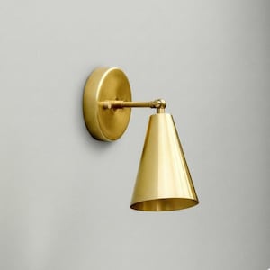 Op de afbeelding: Een wandlamp met een ronde messing muurplaat en een conische messing kap. De lamp is voorzien van een verstelbare arm met een draaibaar gewricht dat de kap met de basis verbindt, uitgevoerd in een gepolijste metallic gouden kleur.