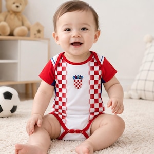 Puede incluir: Un body de bebé de manga corta con un panel central blanco y un patrón de cuadros rojos y blancos en los laterales y las mangas. La prenda muestra el escudo de armas de Croacia en el pecho y tiene ribetes rojos en el cuello y las aberturas de las piernas.