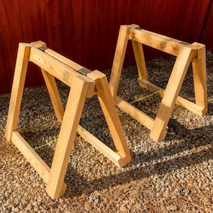 May include: Two light-colored wooden sawhorses with an A-frame design. Each sawhorse features four angled legs connected by horizontal support beams and a top rail. The wood grain is visible throughout the structure, and metal hardware is present.