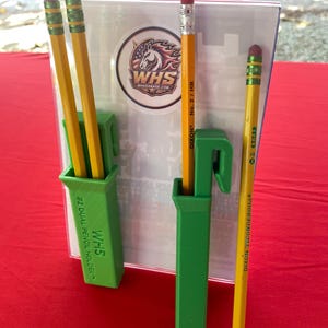 May include: Two green plastic pencil holders with hooks, each featuring the text WHS #2 DUAL PENCIL HOLDER. The holders contain yellow Dixon Ticonderoga pencils with pink erasers. A circular logo with a horse and the text WHS is visible behind them.