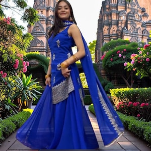 Puede incluir: Un vibrante atuendo tradicional azul con intrincados bordados plateados. El conjunto incluye una blusa sin mangas, una falda fluida y una bufanda a juego. Brazaletes dorados añaden un toque de elegancia. El atuendo se presenta sobre un fondo de exuberante vegetación y un templo.