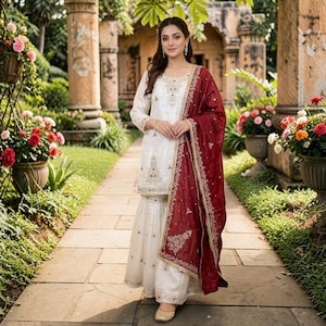 Peut inclure: Un ensemble salwar kameez blanc brodé avec un dupatta bordeaux. La tenue présente des détails dorés complexes et est portée avec des chaussures dorées. L'arrière-plan comprend un sentier en pierre et de la verdure.