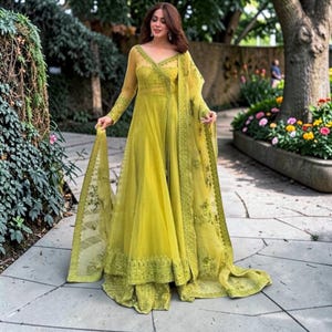 May include: A long, flowing chartreuse dress with a sheer overlay and intricate embroidery. The dress features long sleeves and a wrap-style bodice. A matching scarf is draped over the shoulder. The outfit is worn outdoors.