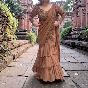 May include: A brown saree with a tiered, ruffled skirt and a long-sleeved, embellished top. The saree features intricate gold detailing along the edges and bodice. The fabric drapes elegantly, creating a flowing silhouette. The saree is worn outdoors.