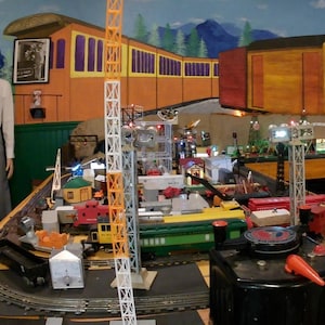 Könnte beinhalten: Eine detaillierte Modelleisenbahnanlage mit einem hohen, orange-weiß karierten Turm. Die Szene umfasst Miniaturzüge, Gebäude und ein Bedienfeld, vor dem Hintergrund einer gemalten Landschaft mit einem Waggon.
