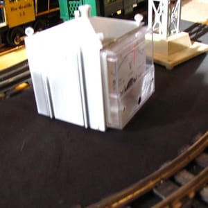 May include: A white model train control box with a clear front panel displaying a gauge. The box is rectangular with a ridged design on the side. The scene includes model train tracks and a yellow and black train car.