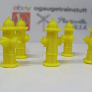 Op de afbeelding: Een verzameling van zes felgele miniatuurbrandkranen. Elke brandkraan heeft een afgeronde bovenkant, een cilindrisch lichaam en verschillende uitstekende sproeiers. De brandkranen staan op een witte ondergrond.
