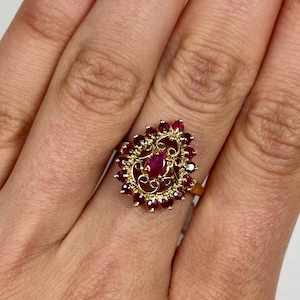 May include: A gold ring featuring a pear-shaped central ruby surrounded by a halo of smaller round rubies. The setting includes intricate gold filigree scrollwork, creating a teardrop-shaped cluster design that sits prominently on the finger.
