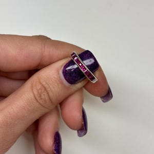 May include: A silver-toned band ring featuring a channel setting with a row of seven square-cut, vibrant magenta-pink gemstones. The ring is positioned horizontally across a fingernail, showcasing the metallic band and the faceted stones.
