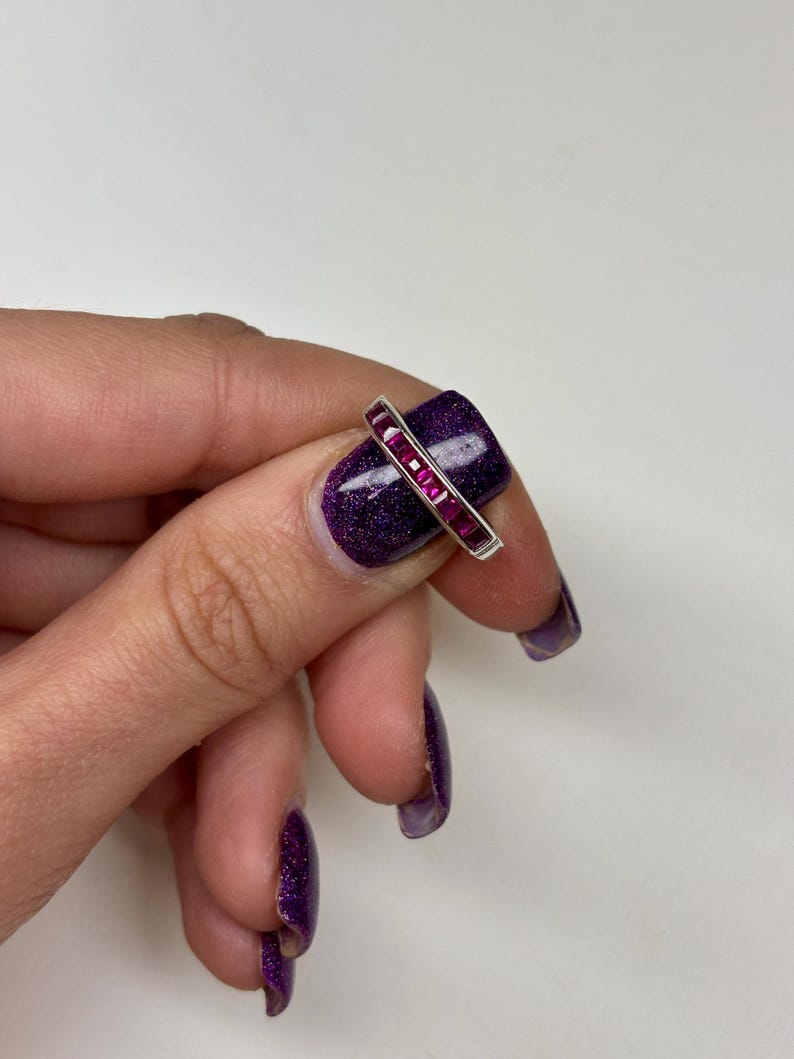 May include: A silver-toned band ring featuring a channel setting with a row of seven square-cut, vibrant magenta-pink gemstones. The ring is positioned horizontally across a fingernail, showcasing the metallic band and the faceted stones.