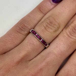 May include: A silver-toned band ring featuring a channel setting with a row of seven square-cut, deep pinkish-red gemstones. The stones are aligned horizontally along the center of the metal band, creating a continuous, uniform appearance.