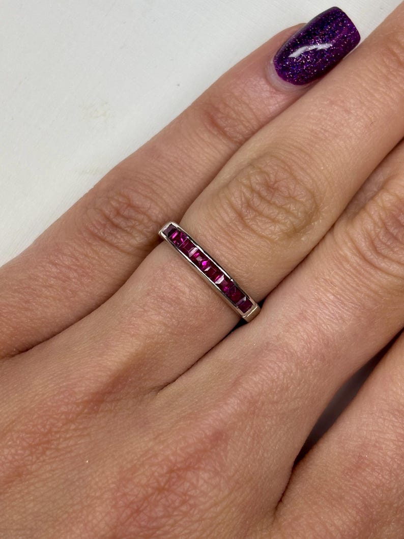 May include: A silver-toned band ring featuring a channel setting with a row of seven square-cut, deep pinkish-red gemstones. The stones are aligned horizontally along the center of the metal band, creating a continuous, uniform appearance.