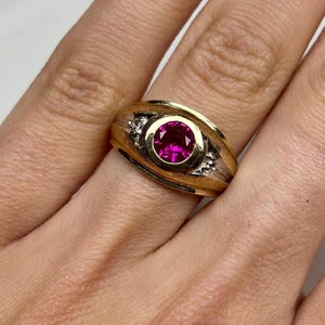 May include: A gold ring featuring a round, vibrant pink gemstone set in a bezel at the center. The band has a textured, brushed finish with two small, clear accent stones embedded on either side of the central setting, creating a detailed look.