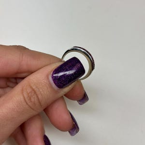 May include: A silver-toned metal ring featuring a curved band with a series of small, dark red, rectangular-cut stones set along the top surface. The ring has a polished finish and a smooth, rounded interior band, showcasing a simple design.