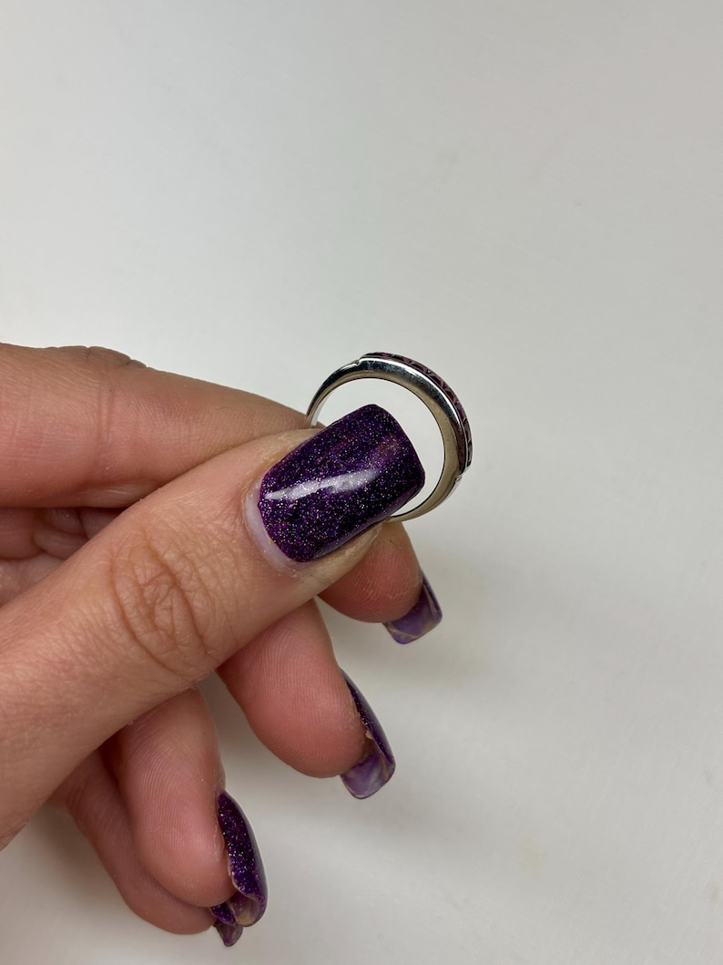May include: A silver-toned metal ring featuring a curved band with a series of small, dark red, rectangular-cut stones set along the top surface. The ring has a polished finish and a smooth, rounded interior band, showcasing a simple design.