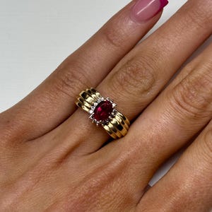 May include: A gold ring featuring a central oval-cut red gemstone surrounded by a halo of small clear stones. The band is constructed from three parallel, rounded gold bands that merge together, creating a wide, textured, and polished appearance.