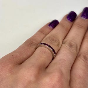 May include: A silver-toned band ring featuring a channel setting with a series of small, square-cut, deep red gemstones embedded along the top half of the band. The ring has a smooth, polished metal finish and a consistent width throughout its design.