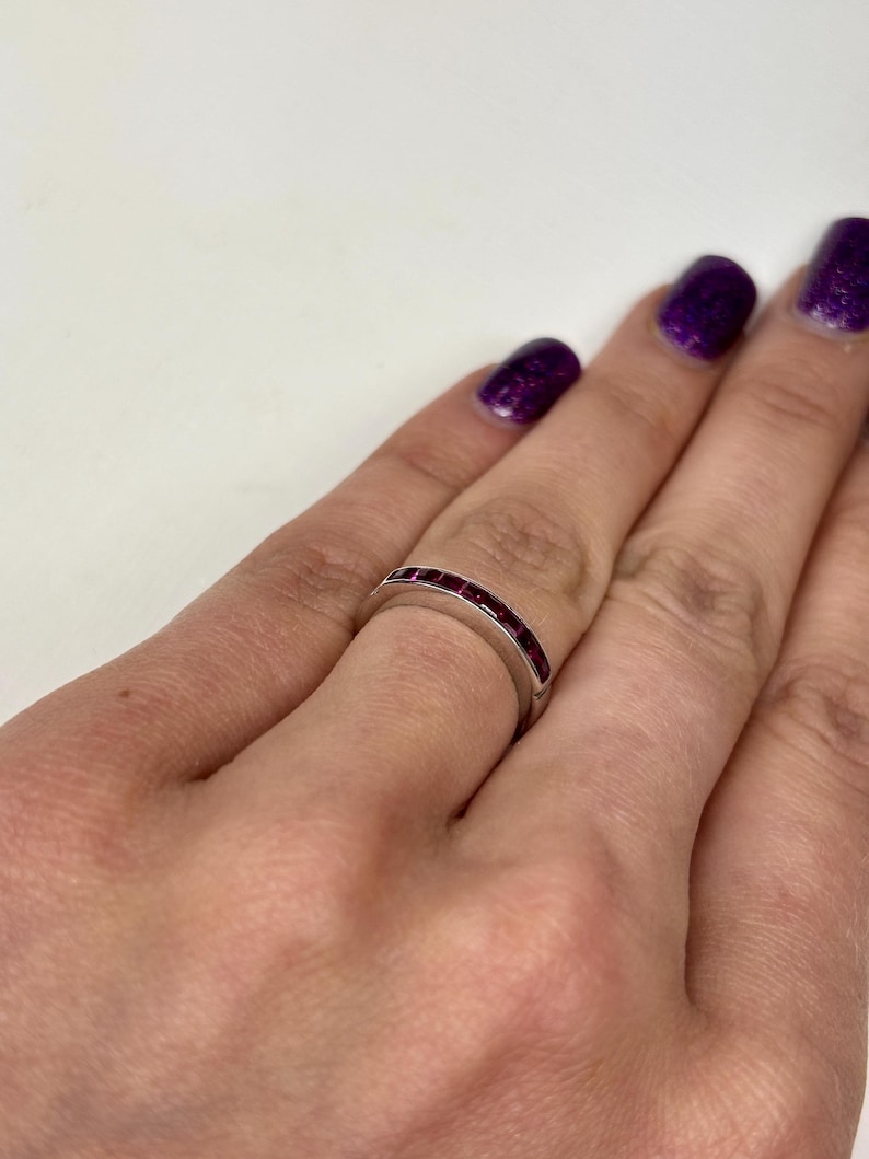 May include: A silver-toned band ring featuring a channel setting with a series of small, square-cut, deep red gemstones embedded along the top half of the band. The ring has a smooth, polished metal finish and a consistent width throughout its design.