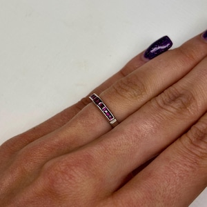 May include: A silver-toned band ring featuring a channel setting with a row of small, rectangular-cut vibrant pink stones. The ring is worn on a finger, showcasing the metallic band and the series of aligned, deep pink gemstones set horizontally.