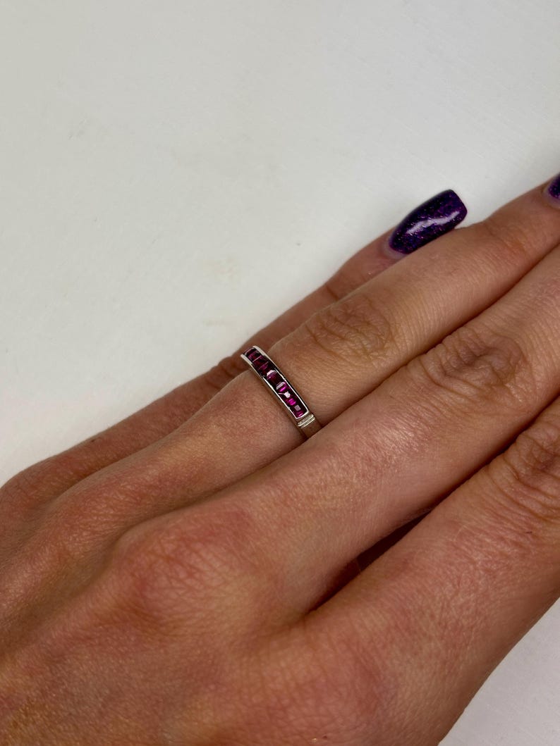 May include: A silver-toned band ring featuring a channel setting with a row of small, rectangular-cut vibrant pink stones. The ring is worn on a finger, showcasing the metallic band and the series of aligned, deep pink gemstones set horizontally.