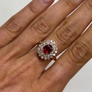 May include: A gold ring featuring a large, round, deep red gemstone surrounded by a halo of smaller, clear gemstones. The ring is set on a gold band and is displayed on a hand.