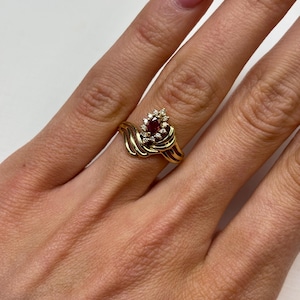 May include: A gold ring featuring a marquise-cut red gemstone at the center, surrounded by a cluster of small round white stones. The band has a bypass design with three parallel gold ribbons that taper toward the back of the ring.