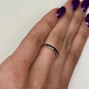 May include: A silver-toned metal band ring featuring a channel setting with a series of small, square-cut deep red gemstones embedded along the top half of the band. The ring has a smooth, polished finish and a consistent width throughout its design.