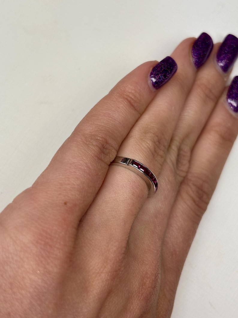 May include: A silver-toned metal band ring featuring a channel setting with a series of small, square-cut deep red gemstones embedded along the top half of the band. The ring has a smooth, polished finish and a consistent width throughout its design.
