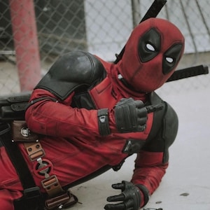 Puede incluir: Una persona con un disfraz de Deadpool rojo y negro, arrodillada y se&ntilde;alando con el dedo. El disfraz incluye una m&aacute;scara con ojos blancos, guantes negros y un cintur&oacute;n de herramientas. Dos espadas est&aacute;n atadas a la espalda.