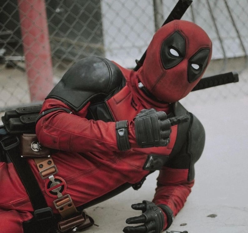 Puede incluir: Una persona con un disfraz de Deadpool rojo y negro, arrodillada y se&ntilde;alando con el dedo. El disfraz incluye una m&aacute;scara con ojos blancos, guantes negros y un cintur&oacute;n de herramientas. Dos espadas est&aacute;n atadas a la espalda.