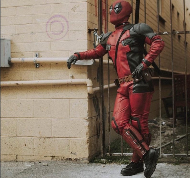 Puede incluir: Un disfraz de Deadpool rojo y negro, que incluye una m&aacute;scara, guantes y botas. El disfraz presenta una combinaci&oacute;n de colores rojo y negro con un cintur&oacute;n de herramientas y una espada en la espalda. El personaje est&aacute; apoyado en una pared de ladrillos y una puerta de metal.
