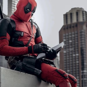 Puede incluir: Una persona con un disfraz de Deadpool rojo y negro, con m&aacute;scara, est&aacute; sentada al aire libre, escribiendo en un cuaderno. El disfraz presenta guantes y detalles en negro. Un paisaje urbano con un edificio alto en el fondo.