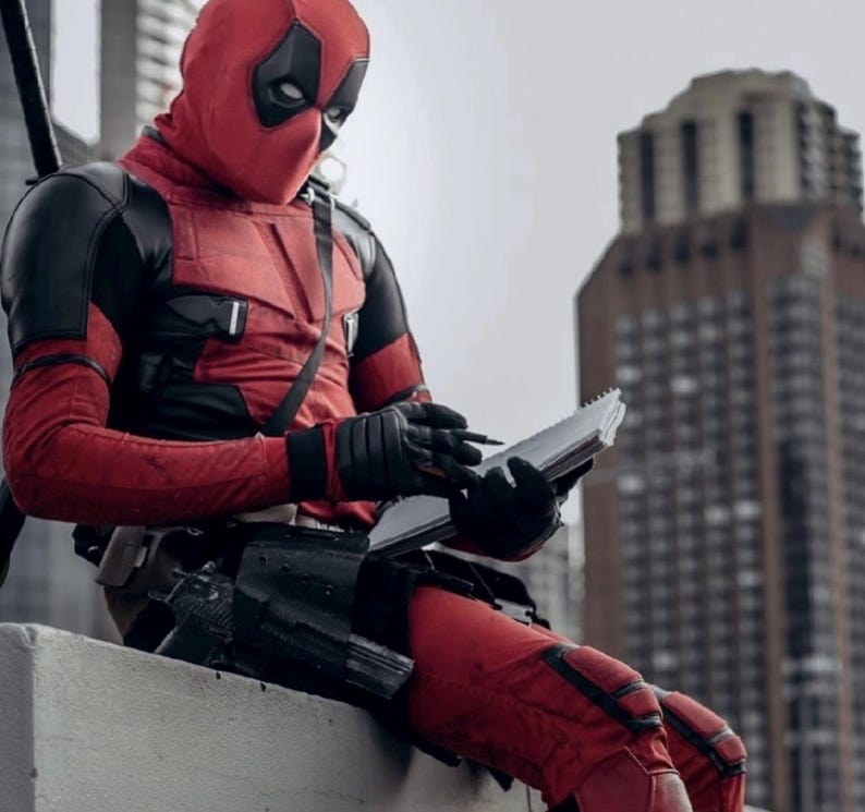 Puede incluir: Una persona con un disfraz de Deadpool rojo y negro, con m&aacute;scara, est&aacute; sentada al aire libre, escribiendo en un cuaderno. El disfraz presenta guantes y detalles en negro. Un paisaje urbano con un edificio alto en el fondo.