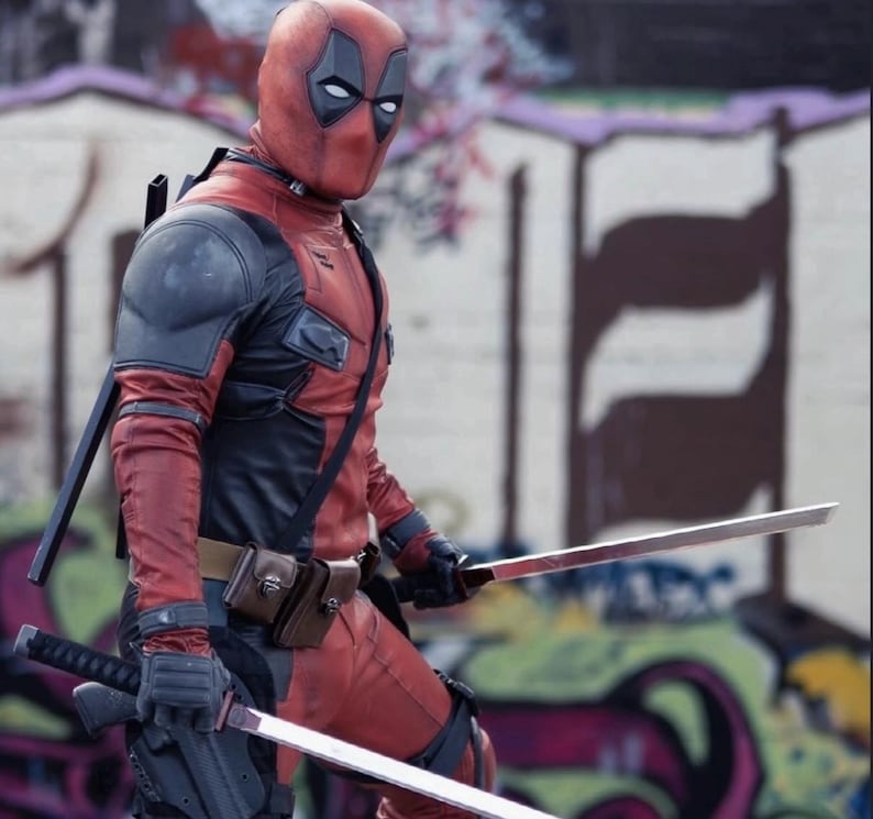 Puede incluir: Una persona con un disfraz de Deadpool rojo y negro, con una m&aacute;scara, sosteniendo dos espadas. El disfraz tiene detalles en negro en los hombros y el pecho, con un cintur&oacute;n de herramientas y bolsas. El fondo presenta arte de graffiti.