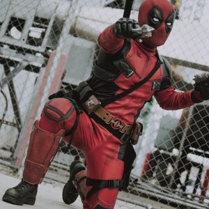 Puede incluir: Una persona con un disfraz de Deadpool, arrodillada y apuntando con dos pistolas de juguete plateadas. El disfraz es rojo y negro, con una m&aacute;scara que cubre la cara. El fondo incluye una valla de alambre y elementos industriales.