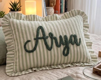 Puede incluir: Un cojín rectangular con un patrón de rayas verticales en verde claro y blanco con un borde con volantes. El nombre Arya está bordado con hilo verde oscuro en letra cursiva en el centro de la superficie del cojín.