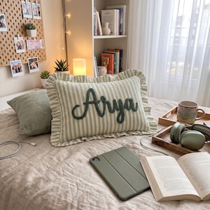 a cozy bedroom scene with a bed, a pillow with the name "Arya" embroidered on it, a laptop, a book, and some other personal items.