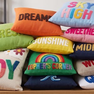 A collection of colorful throw pillows with various words and phrases printed on them, arranged on a wooden surface.