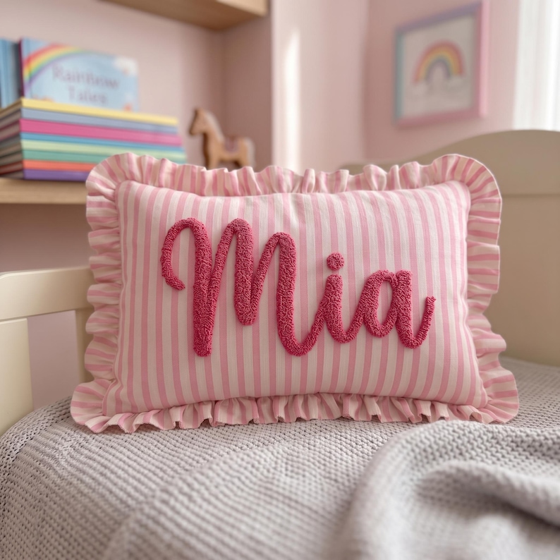 A pink and white striped pillow with the name "Mia" embroidered on it, placed on a bed.