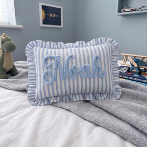A blue and white striped pillow with the name "Noah" written on it, placed on a bed with a gray blanket. In the background, there is a stuffed dinosaur toy and a shelf with various toys.