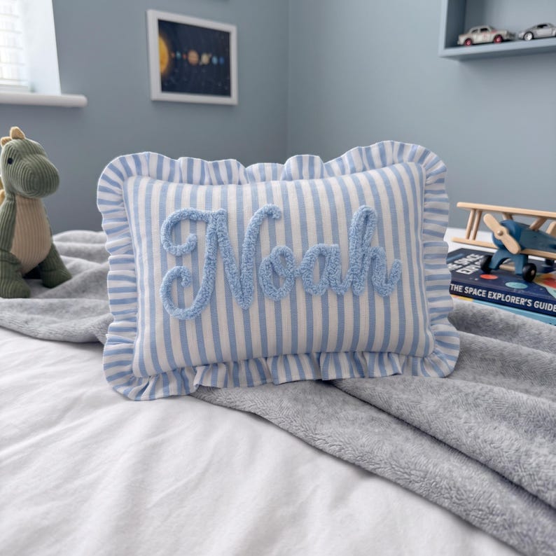 A blue and white striped pillow with the name "Noah" written on it, placed on a bed with a gray blanket. In the background, there is a stuffed dinosaur toy and a shelf with various toys.