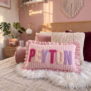 A bedroom with a pink and white striped pillow that says "Peyton" in pink and purple letters, along with other pillows and decorative items.